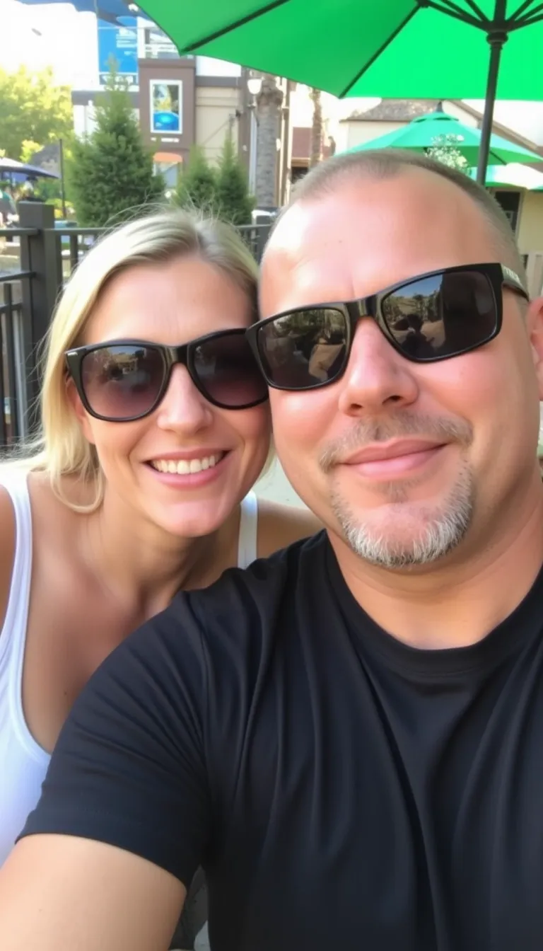 Selfie of a man and woman in their early 50s, both wearing sunglasses, on an outdoor patio with green umbrellas. She is smiling at the camera, while he has short hair, a clean-shaven face, and a goatee. He is wearing a black t-shirt and a white tank top, with a restaurant background