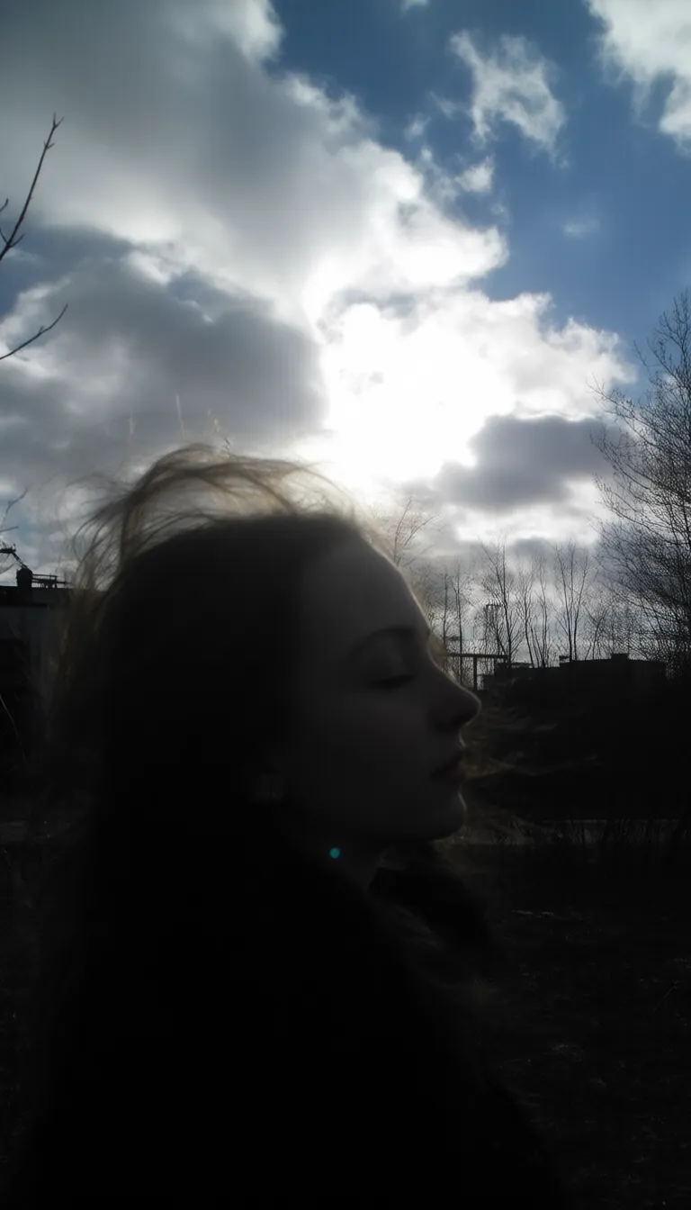 underexposed outdoor scene, raw unedited amateurish candid photo of caucasian young woman, flowing tousled hair, from side view angle, looking at the viewer, dreamy expression, closed eyes, bare winter trees, dramatic cloudy sky, bright sunburst through clouds, deep blue sky patches, industrial urban setting, dark shadowed foreground, late afternoon lighting, silhouetted landscape, underexposed ground elements, natural dramatic lighting, high contrast image. amateur quality, candid style