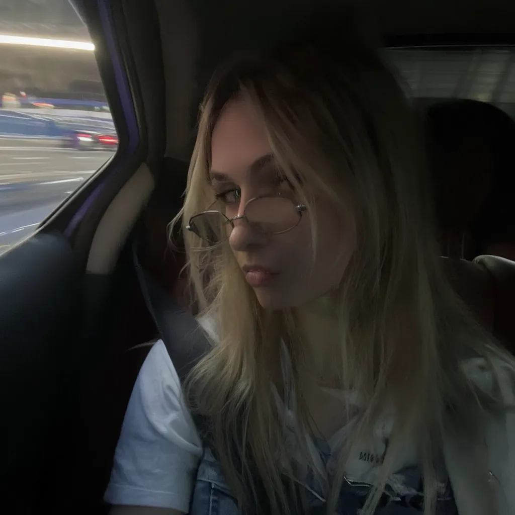 l3n0v0. 20-year-old girl with very pale, smooth skin sitting in the passenger seat of a car, wearing thin wire-rimmed glasses, long blonde hair, slightly wavy, parted in the middle, casual style, wearing a white T-shirt with denim overalls, side profile, looking towards the camera, indoor car setting, seatbelt visible. Background shows motion blur of a highway, streaks of red and white light, suggesting car in motion, shadowed foreground, dark, underexposed, high iso