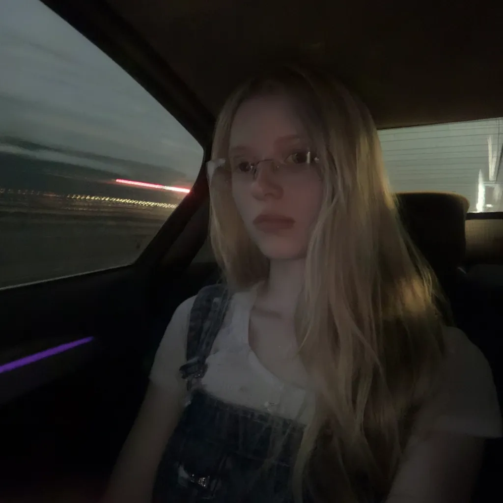 l3n0v0. 20-year-old girl with very pale, smooth skin sitting in the passenger seat of a car, wearing thin wire-rimmed glasses, long blonde hair, slightly wavy, parted in the middle, casual style, wearing a white T-shirt with denim overalls, side profile, looking towards the camera, indoor car setting, seatbelt visible. Background shows motion blur of a highway, streaks of red and white light, suggesting car in motion, shadowed foreground, dark, underexposed, high iso