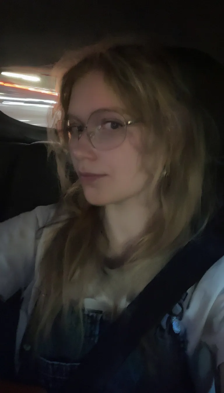 l3n0v0. 20-year-old girl with very pale, smooth skin sitting in the passenger seat of a car, wearing thin wire-rimmed glasses, long blonde hair, slightly wavy, parted in the middle, casual style, wearing a white T-shirt with denim overalls, side profile, looking towards the camera, indoor car setting, seatbelt visible. Background shows motion blur of a highway, streaks of red and white light, suggesting car in motion, shadowed foreground, dark, underexposed, high iso