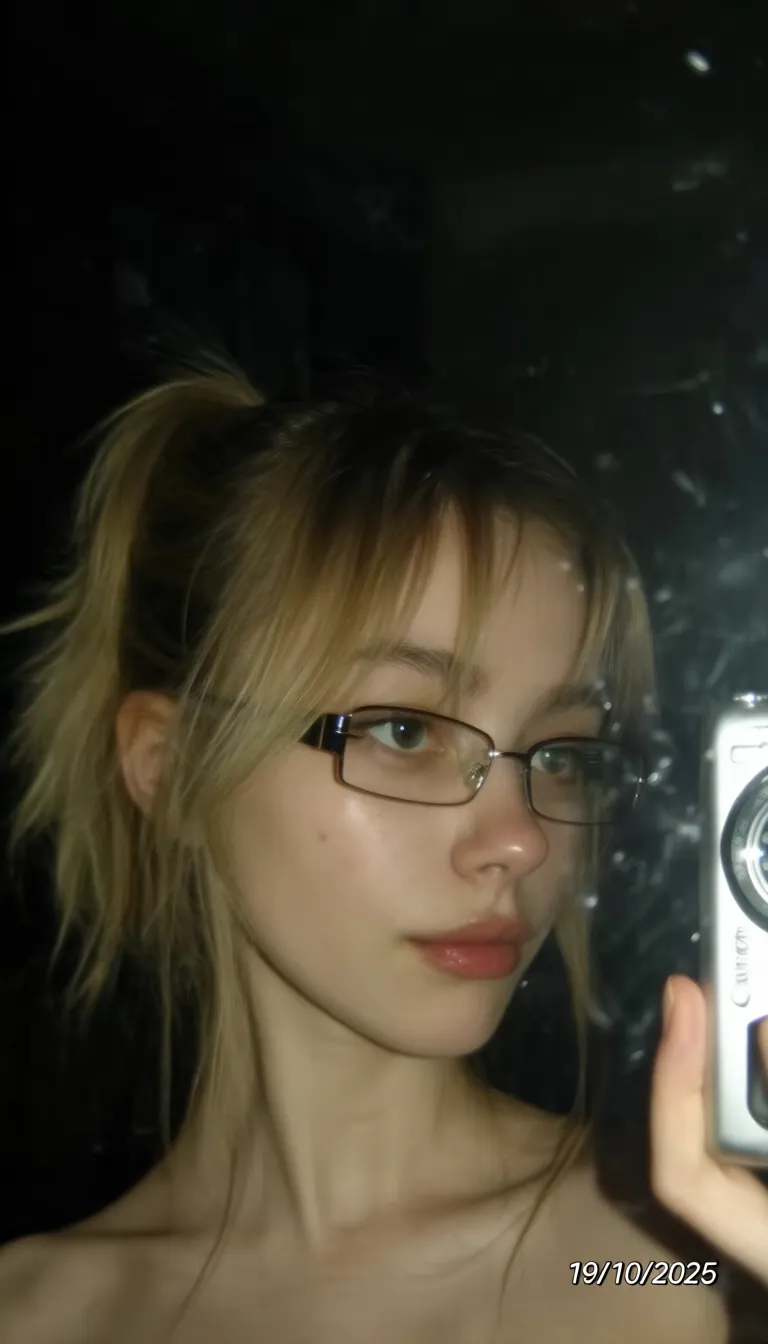 Digital photograph, young 20 years old caucasian woman, she is slim and adorable, goodlit indoor scene, raw unedited amateurish candid photo of selfie, extravant pose, taking photo in mirror with Canon digital camera that she holds in right hand, relaxed expression, gaze directed at camera, stylish 2000s extremely narrow glasses, timestamp overlay "19/10/2025" in bottom right corner, close-up portrait, dim indoor lighting, slightly grainy texture, early 2000s amateur photography, thin small lips, high blonde ponytail with face-framing layers and long bangs, cold tones of photo