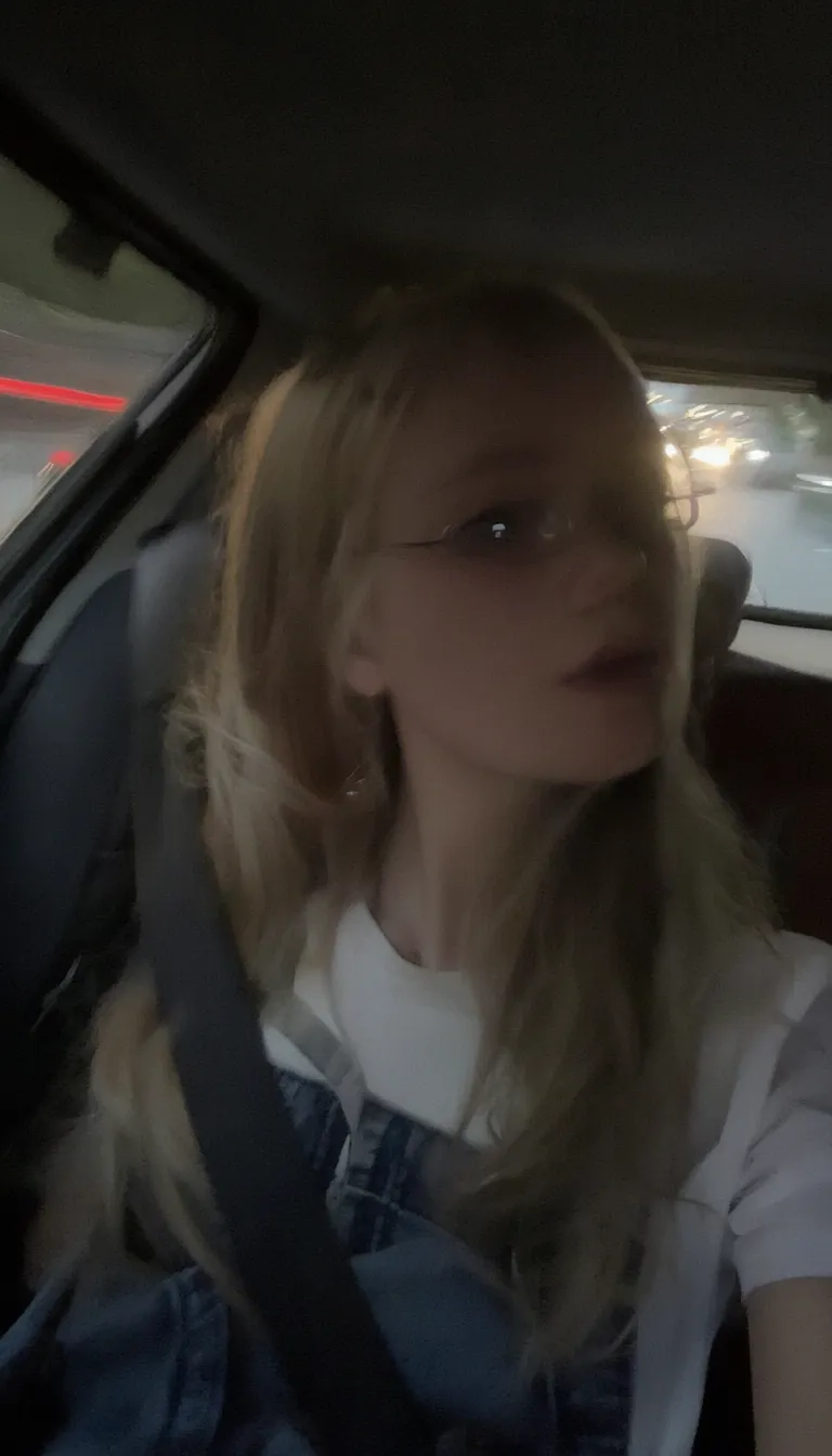 l3n0v0. 20-year-old girl with very pale, smooth skin sitting in the passenger seat of a car, wearing thin wire-rimmed glasses, long blonde hair, slightly wavy, parted in the middle, casual style, wearing a white T-shirt with denim overalls, side profile, looking towards the camera, indoor car setting, seatbelt visible. Background shows motion blur of a highway, streaks of red and white light, suggesting car in motion, shadowed foreground, dark, underexposed, high iso