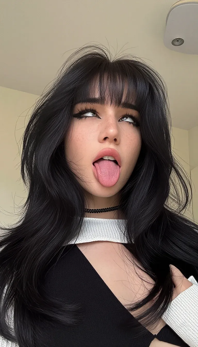ahegao. A medium close-up photograph captures a young woman as the central subject. She is positioned slightly off-center, angled towards the viewer's left, with her head tilted back and her gaze directed upwards and to her right. Her long, voluminous black hair, featuring prominent, straight-cut bangs that partially obscure her forehead, cascades around her face and shoulders. The hair has a wavy texture and appears thick and full.
Her facial features are clearly defined. Her eyes are wide open, with dark, thick eyelashes and light-colored irises visible. Her lips are parted, revealing her upper teeth, and her tongue is extended prominently forward and slightly to her right. She has a light complexion with visible freckles across her nose and cheeks. A thin, black, coiled choker necklace rests at the base of her neck. She is wearing a black and white knitted garment with a high, wide neckline. The white section of the garment is textured with a visible knit pattern and forms a collar-like structure around her neck, while the black fabric covers her shoulders and chest. The garment is open at the front, revealing her collarbones and the top of her chest. Her right shoulder is visible, and her right hand appears to be partially gripping the edge of the garment near her right shoulder. Her left shoulder is mostly obscured by her hair.
The background is an indoor setting with a plain, light-colored wall and ceiling. A modern, cylindrical ceiling light fixture is visible in the upper right corner. The lighting is bright and even, illuminating the subject from the front and slightly above.