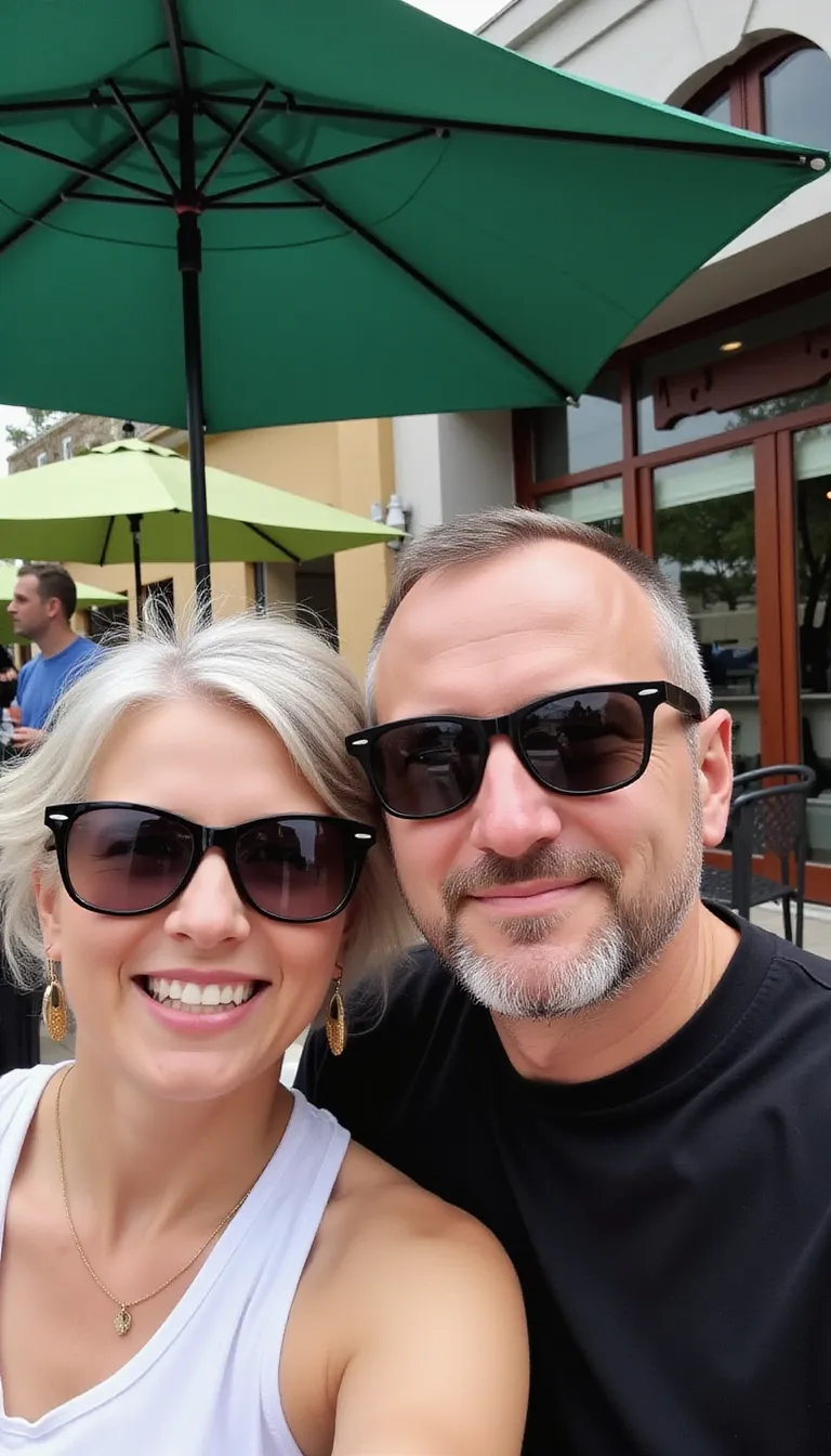 Selfie of a man and woman in their early 50s, both wearing sunglasses, on an outdoor patio with green umbrellas. She is smiling at the camera, while he has short hair, a clean-shaven face, and a goatee. He is wearing a black t-shirt and a white tank top, with a restaurant background