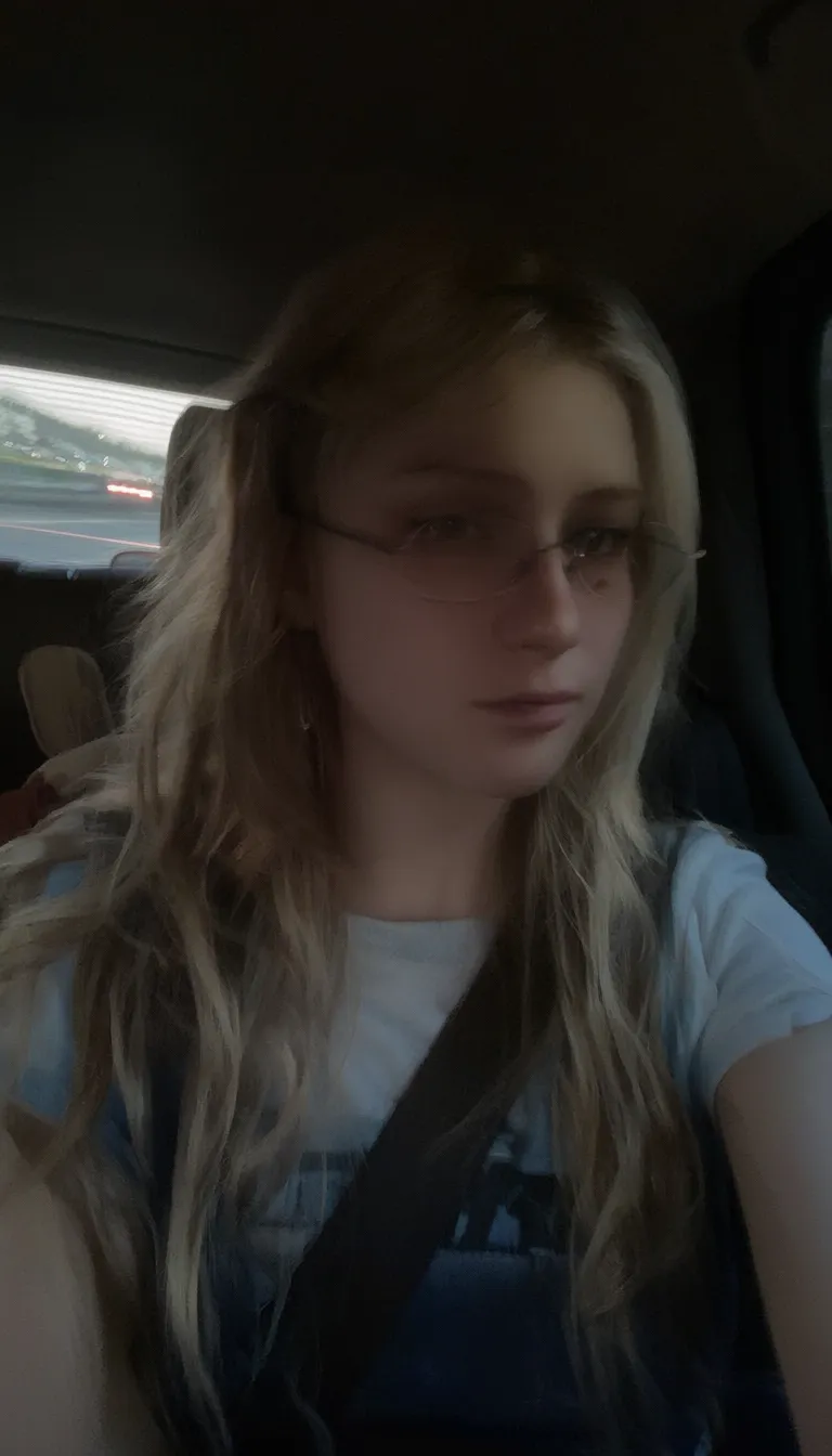 l3n0v0. 20-year-old girl with very pale, smooth skin sitting in the passenger seat of a car, wearing thin wire-rimmed glasses, long blonde hair, slightly wavy, parted in the middle, casual style, wearing a white T-shirt with denim overalls, side profile, looking towards the camera, indoor car setting, seatbelt visible. Background shows motion blur of a highway, streaks of red and white light, suggesting car in motion, shadowed foreground, dark, underexposed, high iso