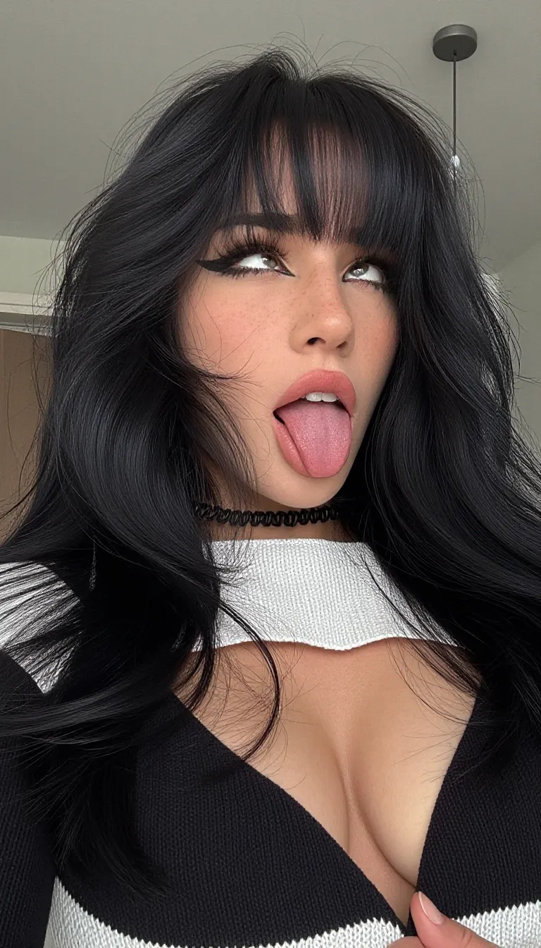 ahegao. A medium close-up photograph captures a young woman as the central subject. She is positioned slightly off-center, angled towards the viewer's left, with her head tilted back and her gaze directed upwards and to her right. Her long, voluminous black hair, featuring prominent, straight-cut bangs that partially obscure her forehead, cascades around her face and shoulders. The hair has a wavy texture and appears thick and full.
Her facial features are clearly defined. Her eyes are wide open, with dark, thick eyelashes and light-colored irises visible. Her lips are parted, revealing her upper teeth, and her tongue is extended prominently forward and slightly to her right. She has a light complexion with visible freckles across her nose and cheeks. A thin, black, coiled choker necklace rests at the base of her neck. She is wearing a black and white knitted garment with a high, wide neckline. The white section of the garment is textured with a visible knit pattern and forms a collar-like structure around her neck, while the black fabric covers her shoulders and chest. The garment is open at the front, revealing her collarbones and the top of her chest. Her right shoulder is visible, and her right hand appears to be partially gripping the edge of the garment near her right shoulder. Her left shoulder is mostly obscured by her hair.
The background is an indoor setting with a plain, light-colored wall and ceiling. A modern, cylindrical ceiling light fixture is visible in the upper right corner. The lighting is bright and even, illuminating the subject from the front and slightly above.